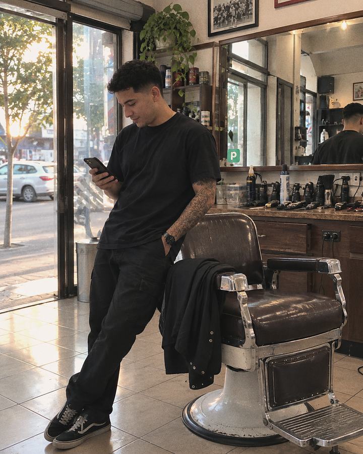 Barbero revisando su teléfono en su barbería, junto al sillón, con la calle al fondo